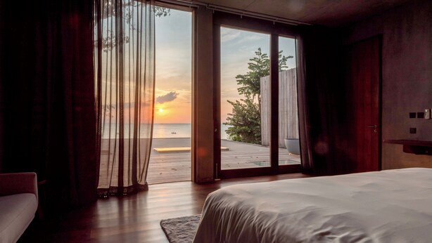 A bedroom with a large bed, a wooden floor, a view of the sunset over the sea, and a sliding glass door leading to an outdoor deck. - Vows Villa Beachfront (Koh Samui)