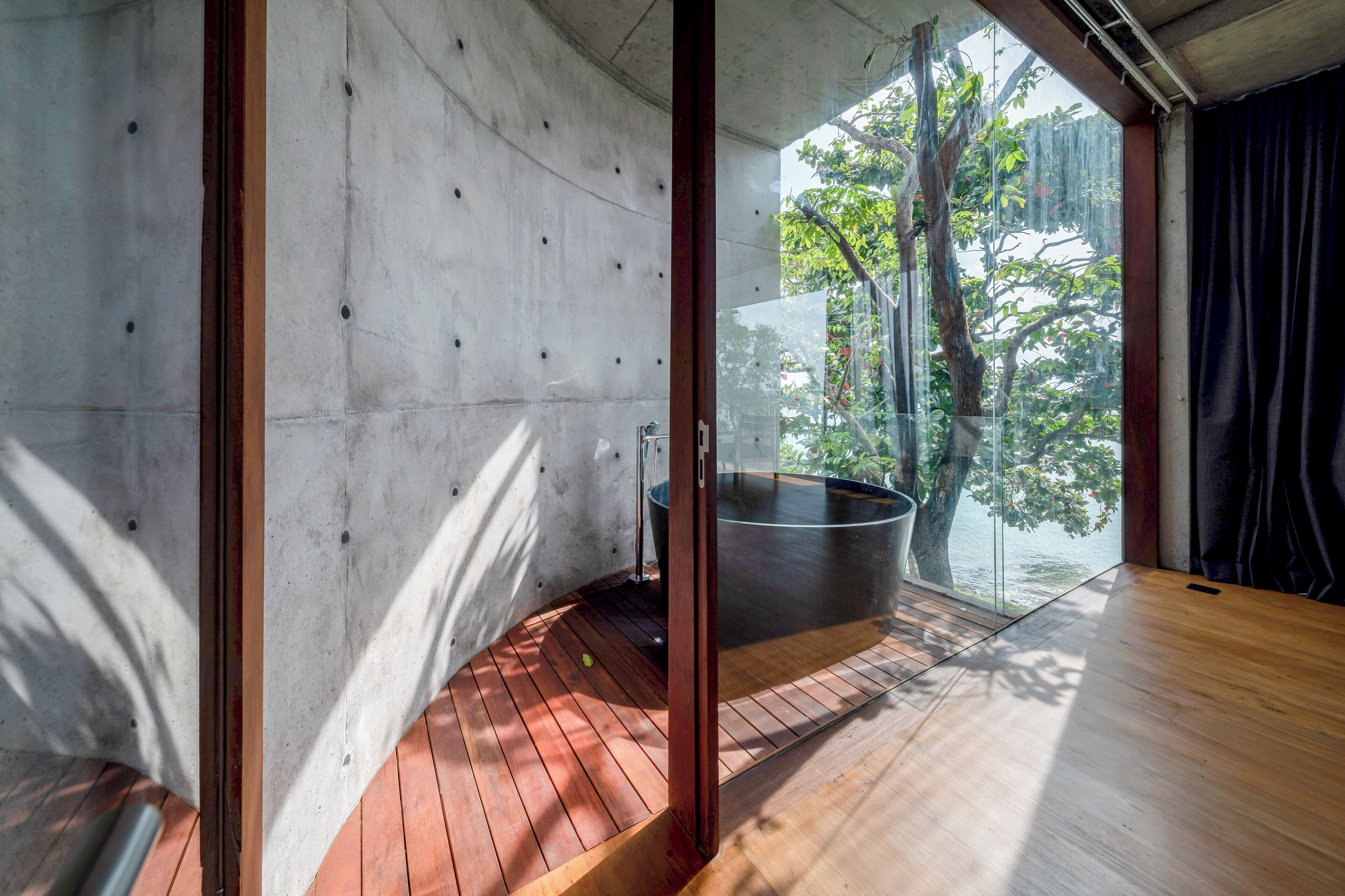 A modern interior with a wooden floor, concrete walls, and a glass door leading to a balcony with a view of trees and a body of water.