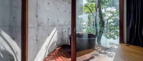 A modern interior with a wooden floor, concrete walls, and a glass door leading to a balcony with a view of trees and a body of water.