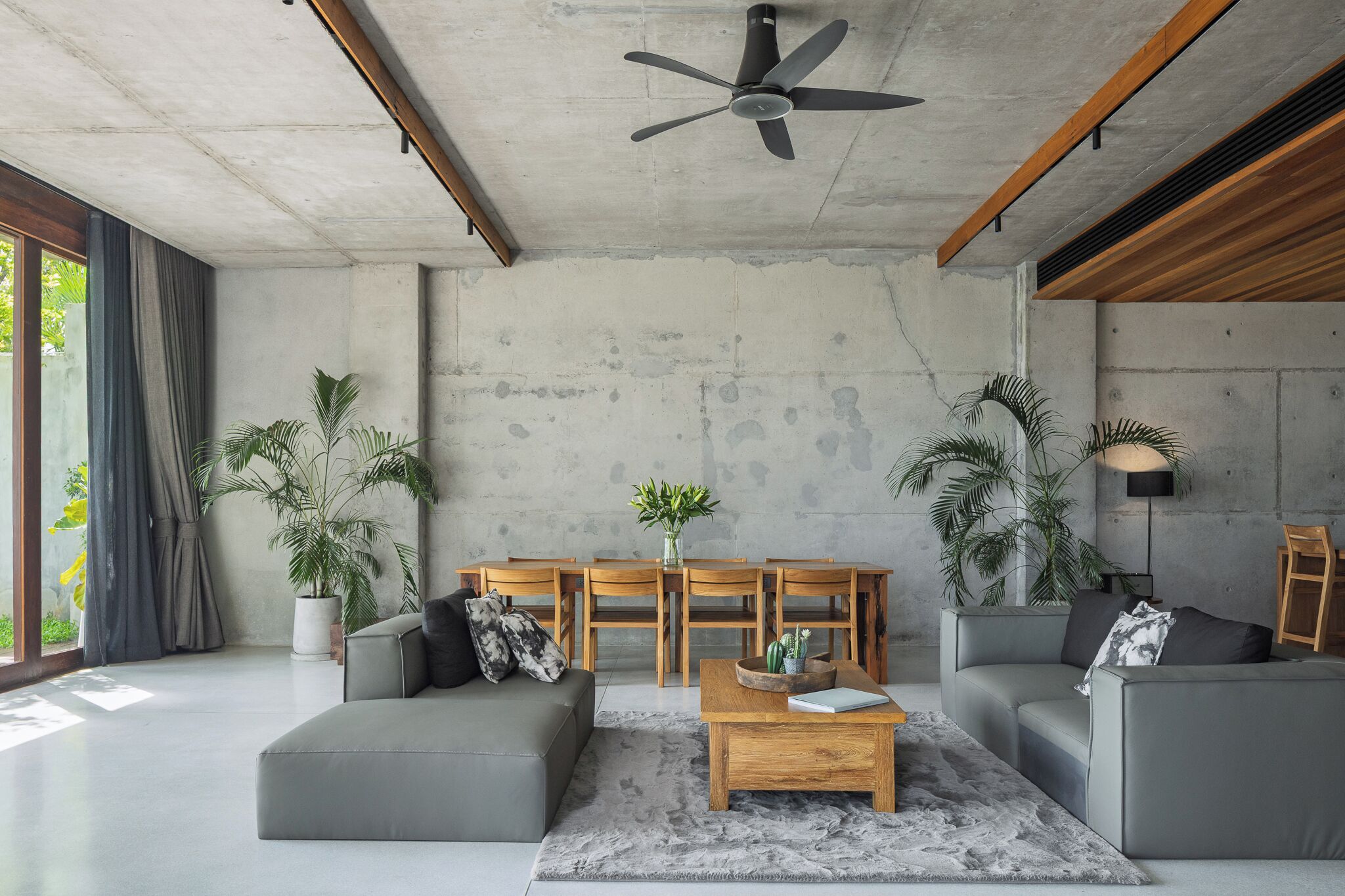 Una sala moderna con un sofá gris, una mesa de centro de madera y un ventilador de techo. Hay plantas en macetas y una mesa de comedor con sillas.