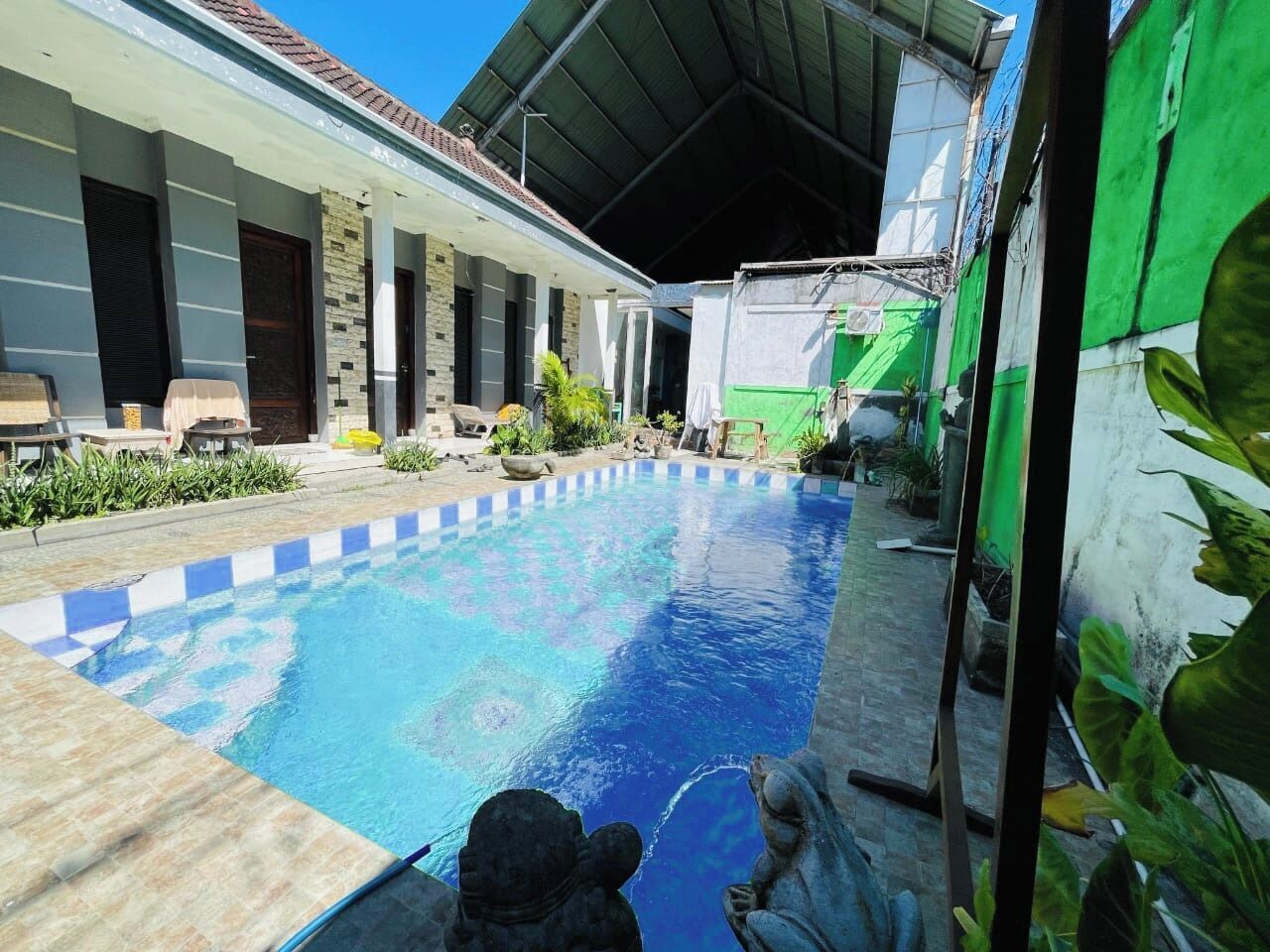 A swimming pool with a tiled deck, surrounded by a covered patio area with outdoor furniture and plants.