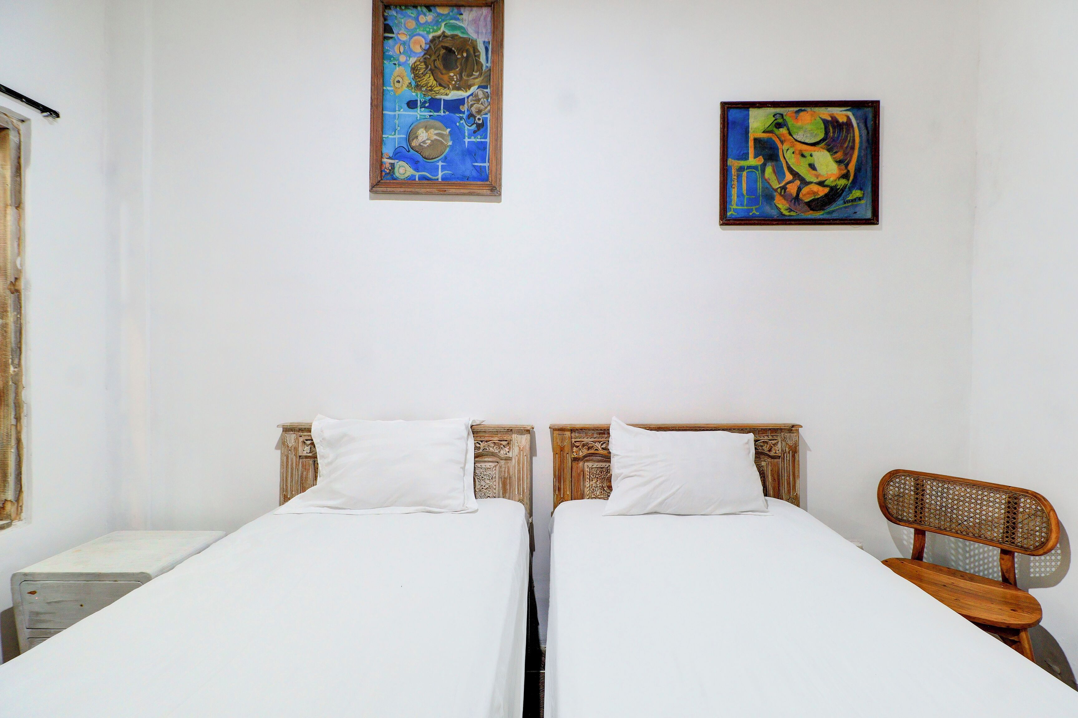 A room with two single beds, a wooden chair, and two paintings on the wall.