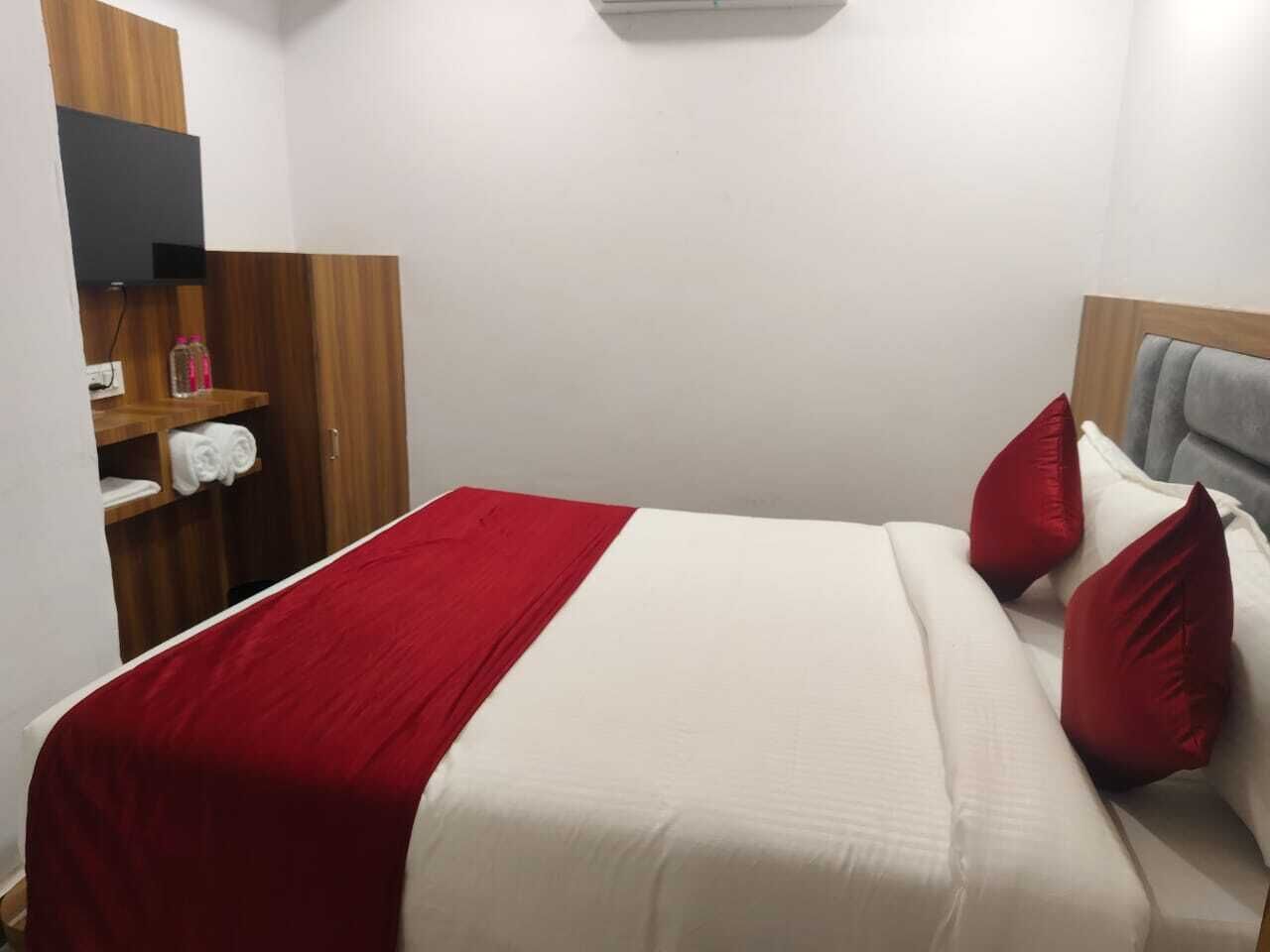 Una cama bien hecha con una colcha roja y blanca, dos almohadas rojas y un cabecero gris. A la izquierda, un armario de madera, y se ve una pequeña estantería con toallas.
