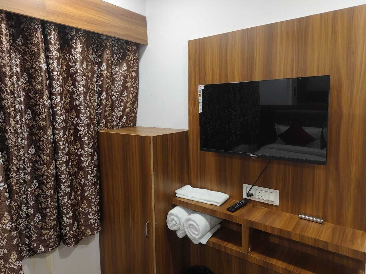 A hotel room with a wooden wardrobe, a flat-screen TV mounted on the wall, a towel rack, and a power outlet.