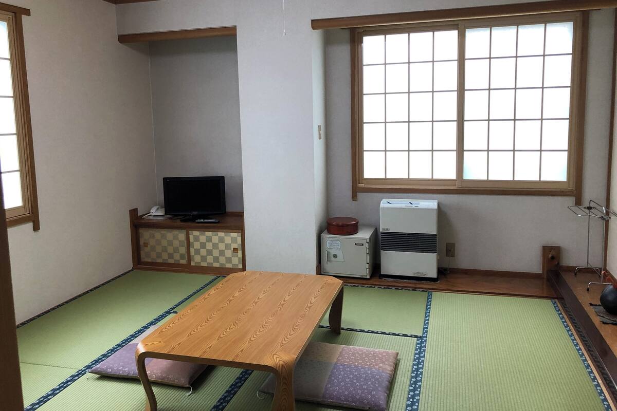 A traditional Japanese room with tatami flooring, a wooden table, a television, and a window with a grid pattern.