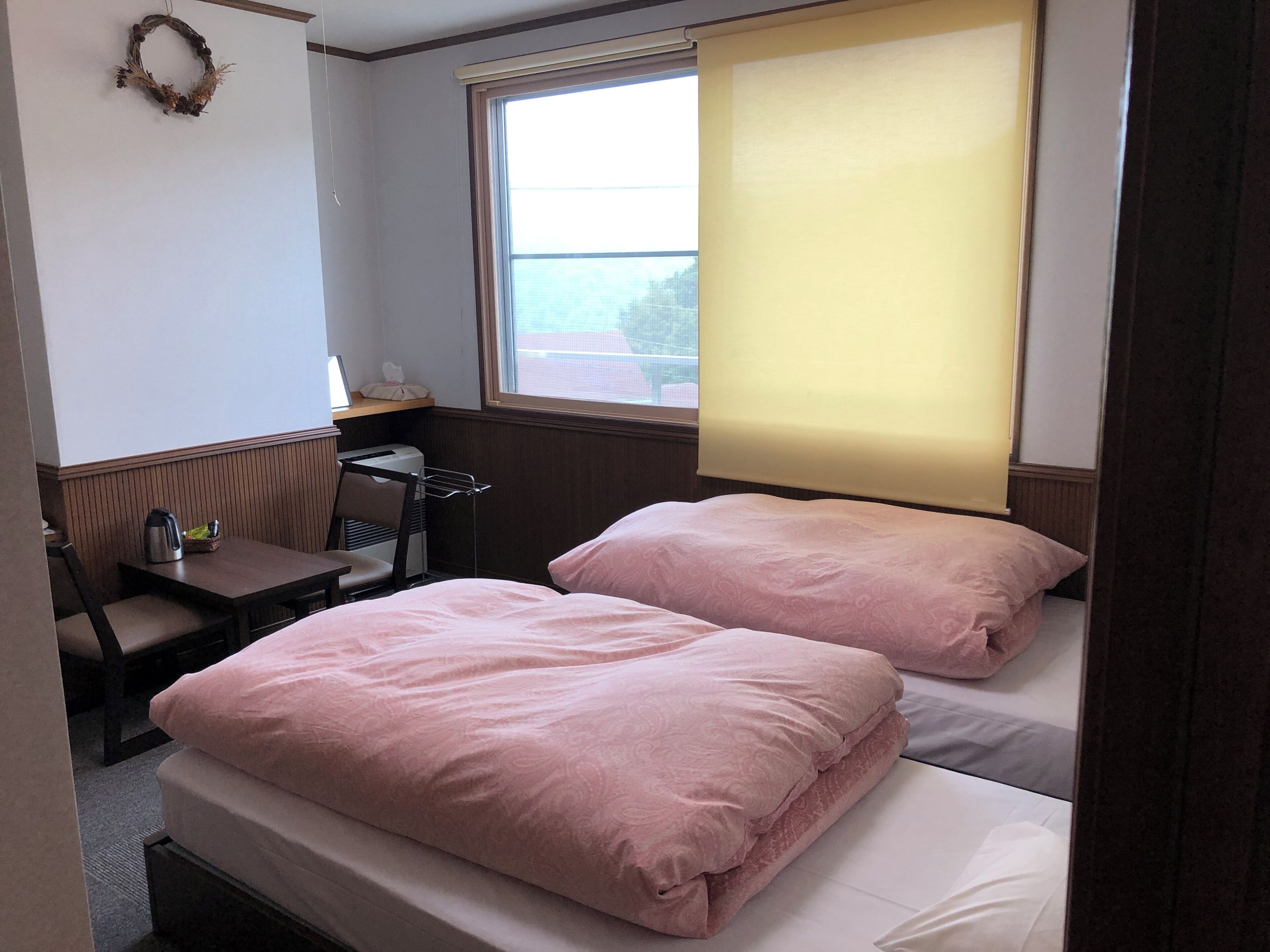 A small, cozy room with two beds, a desk, a chair, and a window with a blind.