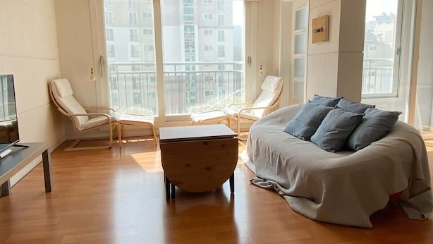 Une chambre d’hôtel moderne, dotée d’un grand lit, de deux fauteuils, d’une petite table et de grandes fenêtres offrant une vue sur les bâtiments.