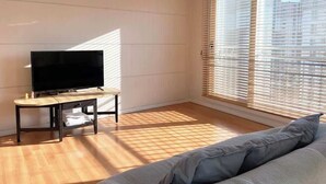 Un salon avec un téléviseur à écran plat posé sur un meuble, un canapé et une fenêtre équipée de stores.