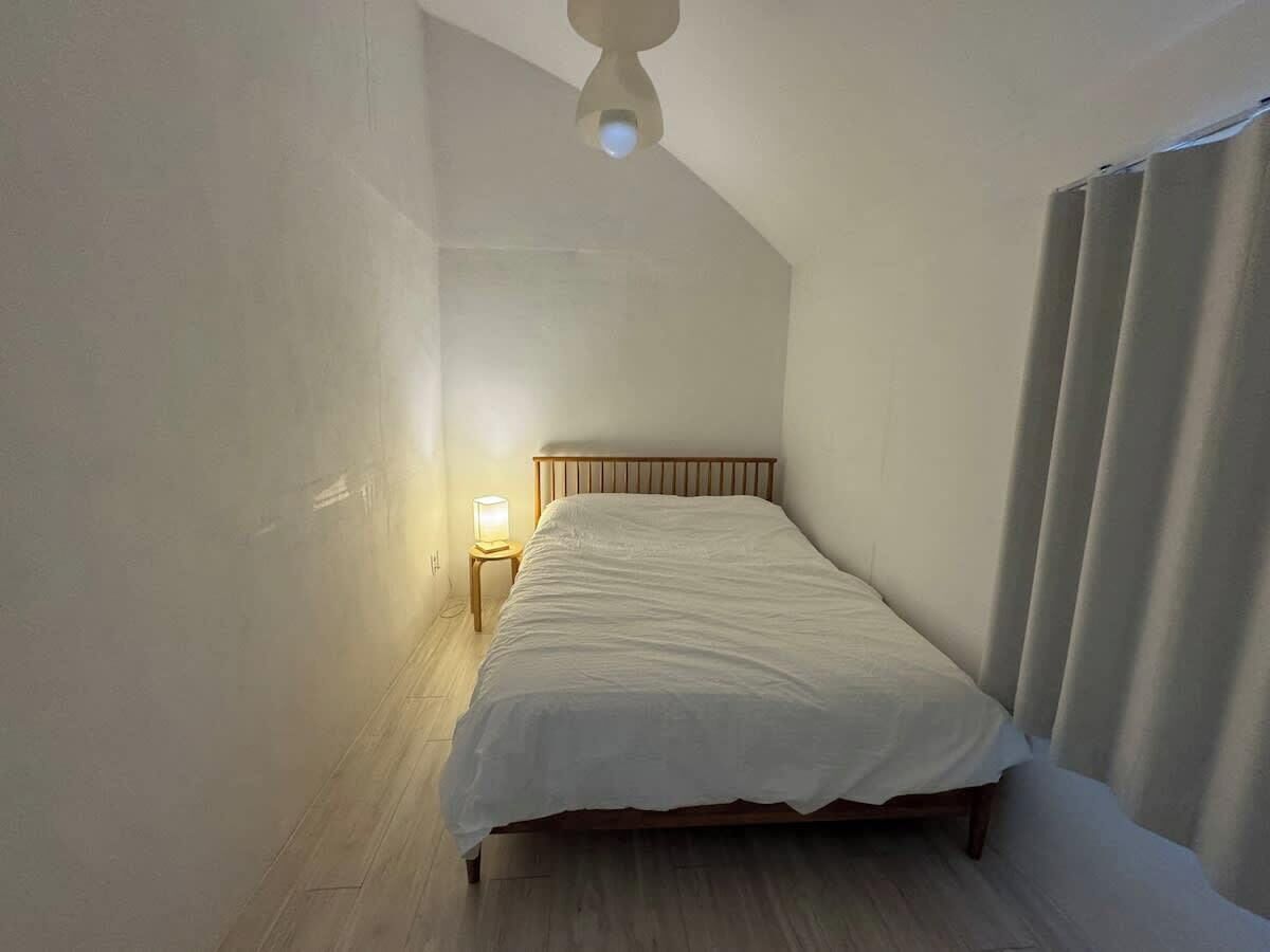 Une chambre à coucher avec un lit, une table de chevet, un luminaire au plafond et une fenêtre avec des rideaux.