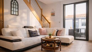 A modern living room with a sofa, a small table with a plant, a staircase, and a sliding glass door to an outdoor area. - Charming house - near train station - 6 people (Les Sables-d´Olonne)