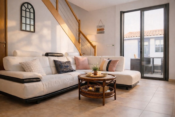 A modern living room with a sofa, a small table with a plant, a staircase, and a sliding glass door to an outdoor area.