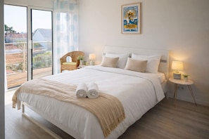 A bedroom with a large bed, a small side table with a lamp, a wicker chair, and a poster on the wall.