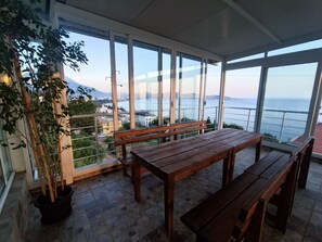 A balcony with a wooden table and benches, large windows offering a view of the sea and cityscape.