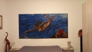 A bedroom with a large painting of a mermaid swimming, a bed with striped bedding, a nightstand, and a wooden sculpture. - Sun Sea Nature b (Sicilia)