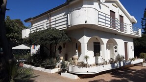 A two-story building with a white facade, multiple balconies, and a garden area with various plants. - Sun Sea Nature b (Sicilia)
