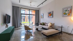 A modern living room with a sofa, ottoman, and chairs, a flat-screen TV, and artwork on the walls. - Swajan Bankers Community (Padra)