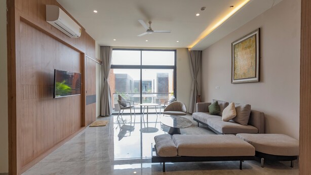 A modern living room with a sofa, ottoman, and a flat-screen TV mounted on the wall. - Swajan Bankers Community (Padra)