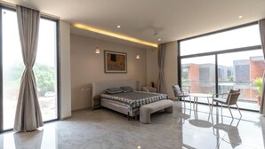 A modern bedroom with a large bed, a small table, and a chair. There is a painting on the wall and a balcony with a view of buildings. - Swajan Bankers Community (Padra)