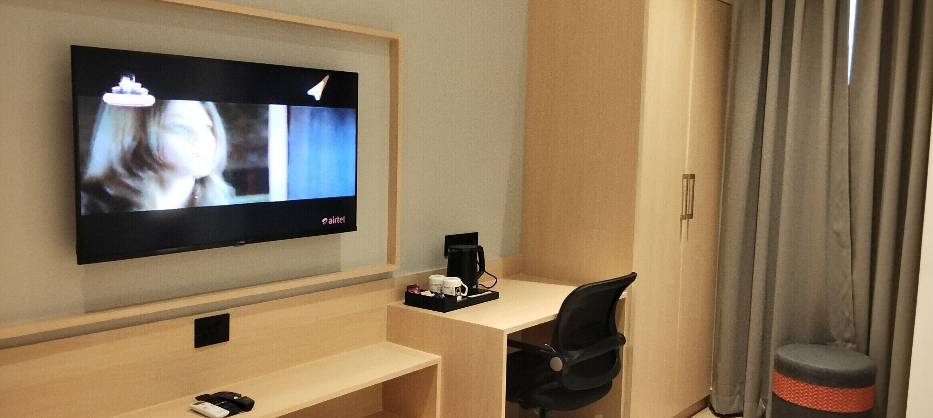 A hotel room with a flat-screen TV mounted on the wall, a wooden desk with a black office chair, a coffee machine, and a small stool.