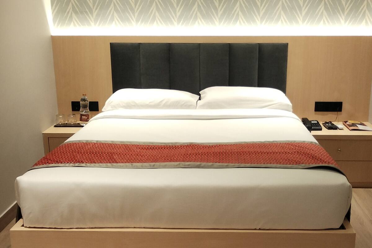 Una cama bien hecha con colcha blanca y roja, cabecera oscura y mesita de noche con lámpara y teléfono.