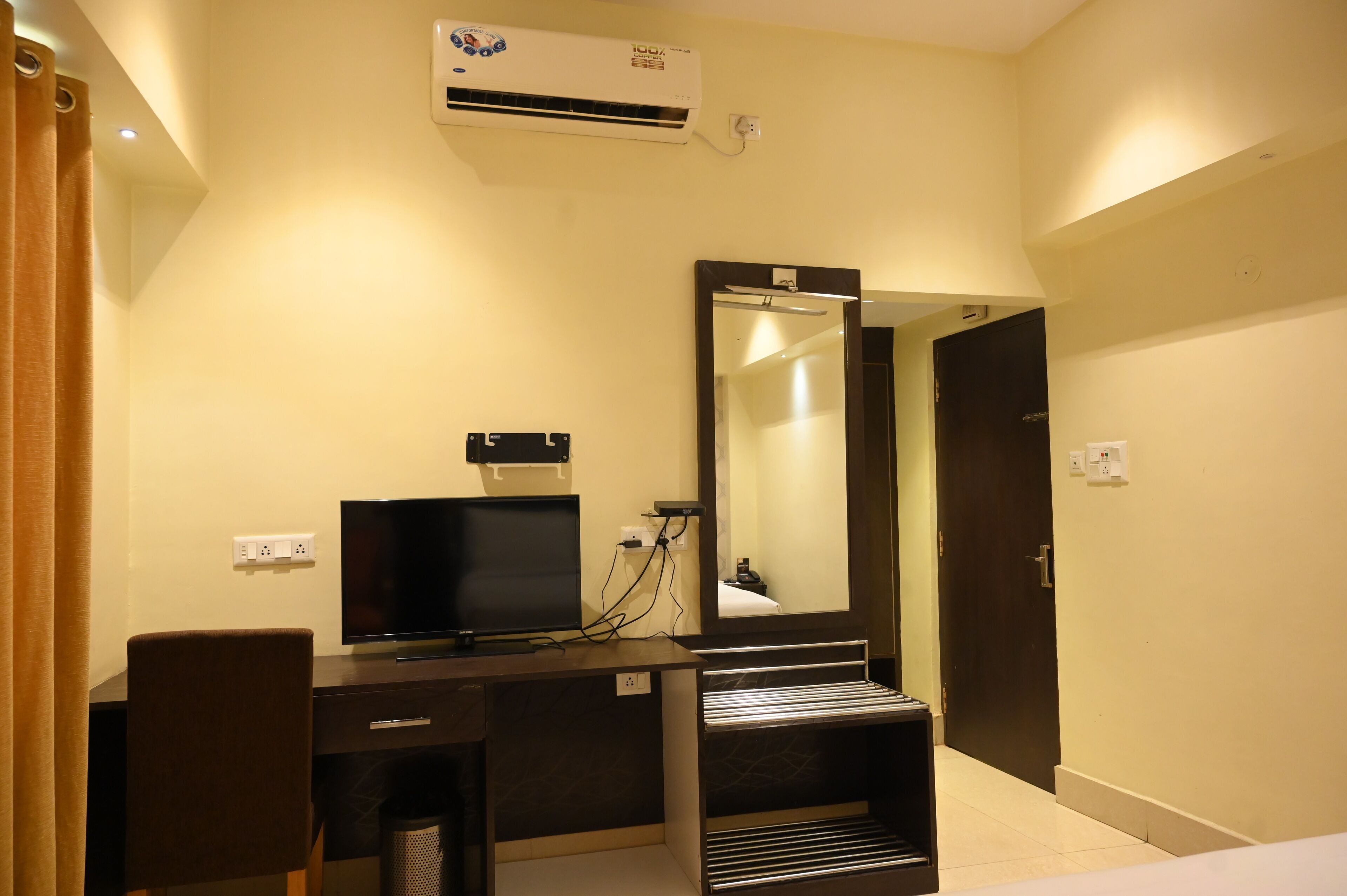 A hotel room with a desk, television, air conditioning unit, mirror, and a door.