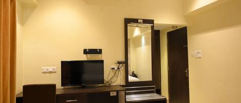 A hotel room with a desk, television, air conditioning unit, mirror, and a door.