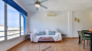 Una sala de estar moderna con un sofá blanco, una mesa de comedor con sillas, un ventilador de techo y un ventanal con vistas a edificios.