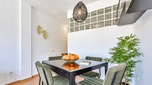 Un comedor con una mesa de vidrio, cuatro sillas tapizadas en verde, un cuenco de fruta, un libro, una lámpara colgante y una planta en maceta.