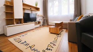 A modern living room with a flat-screen TV, wooden shelves, a sofa, and a window with curtains. - Apartment in the city center (Plovdiv)