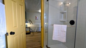 Hotellihuone, jossa on sänky, yöpöytä, lasiseinäinen suihkukaappi ja pyyheteline.
