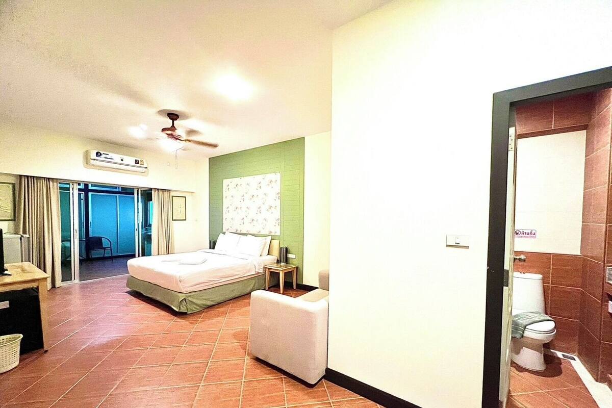 A hotel room with a bed, a small table, a chair, a bedside table, and a bathroom with a toilet and sink.