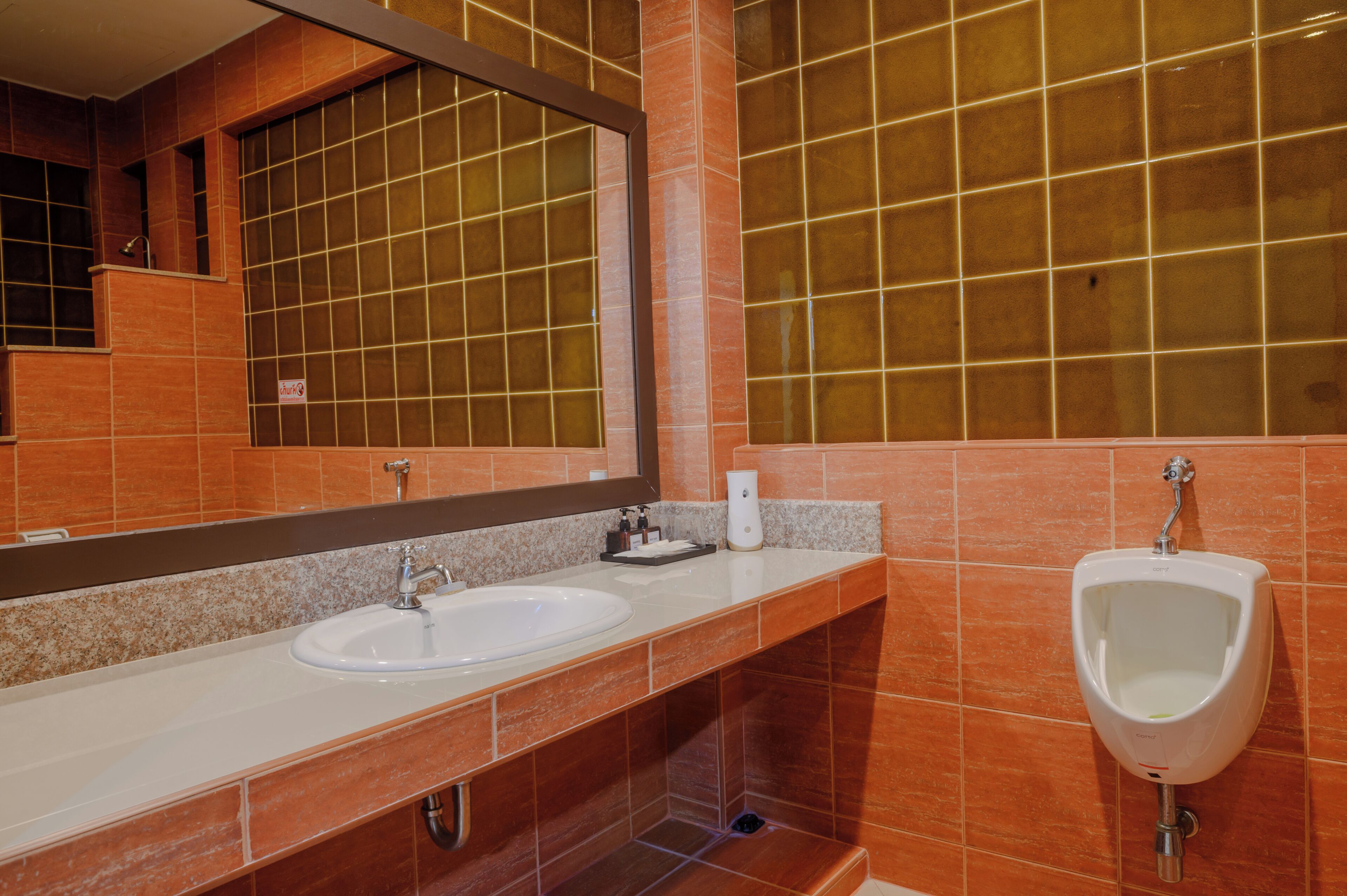 A bathroom with a large mirror, two sinks, a urinal, and orange tiled walls.