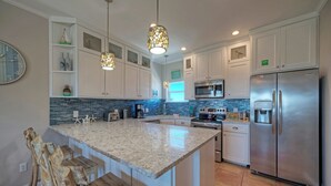 Una cocina moderna con armarios blancos, encimera de granito, refrigerador de acero inoxidable y una lámpara colgante.