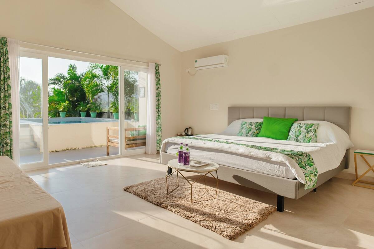 A bedroom with a bed, a table, a bench, and a view of greenery outside.
