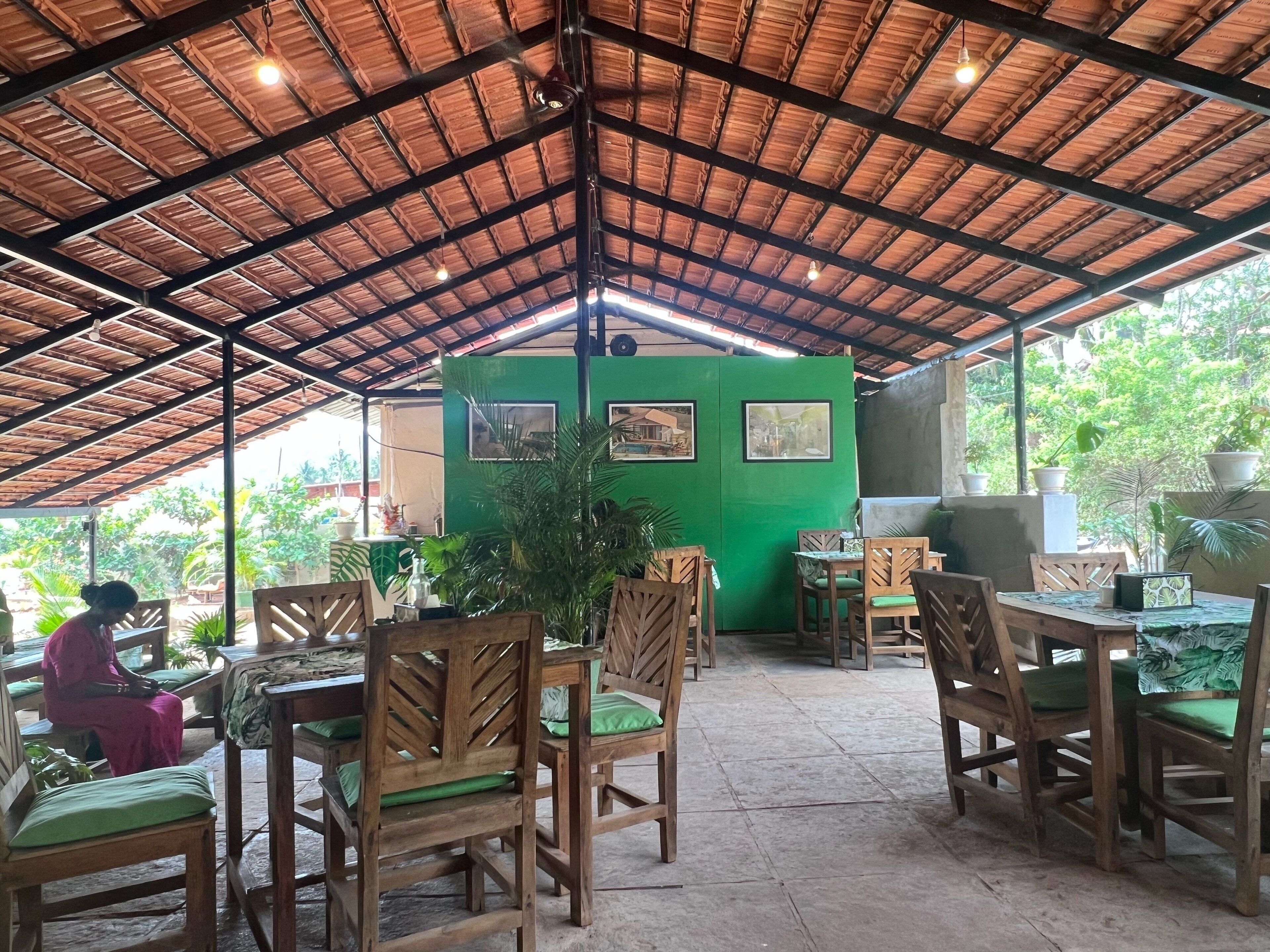 An outdoor cafe with wooden tables and chairs, a green wall with framed pictures, and a thatched roof.
