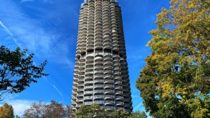 A tall, cylindrical building with numerous balconies, surrounded by trees and a clear blue sky.