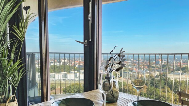 A dining table with two bowls, a wine glass, and a vase with flowers, set against a city view from a balcony.