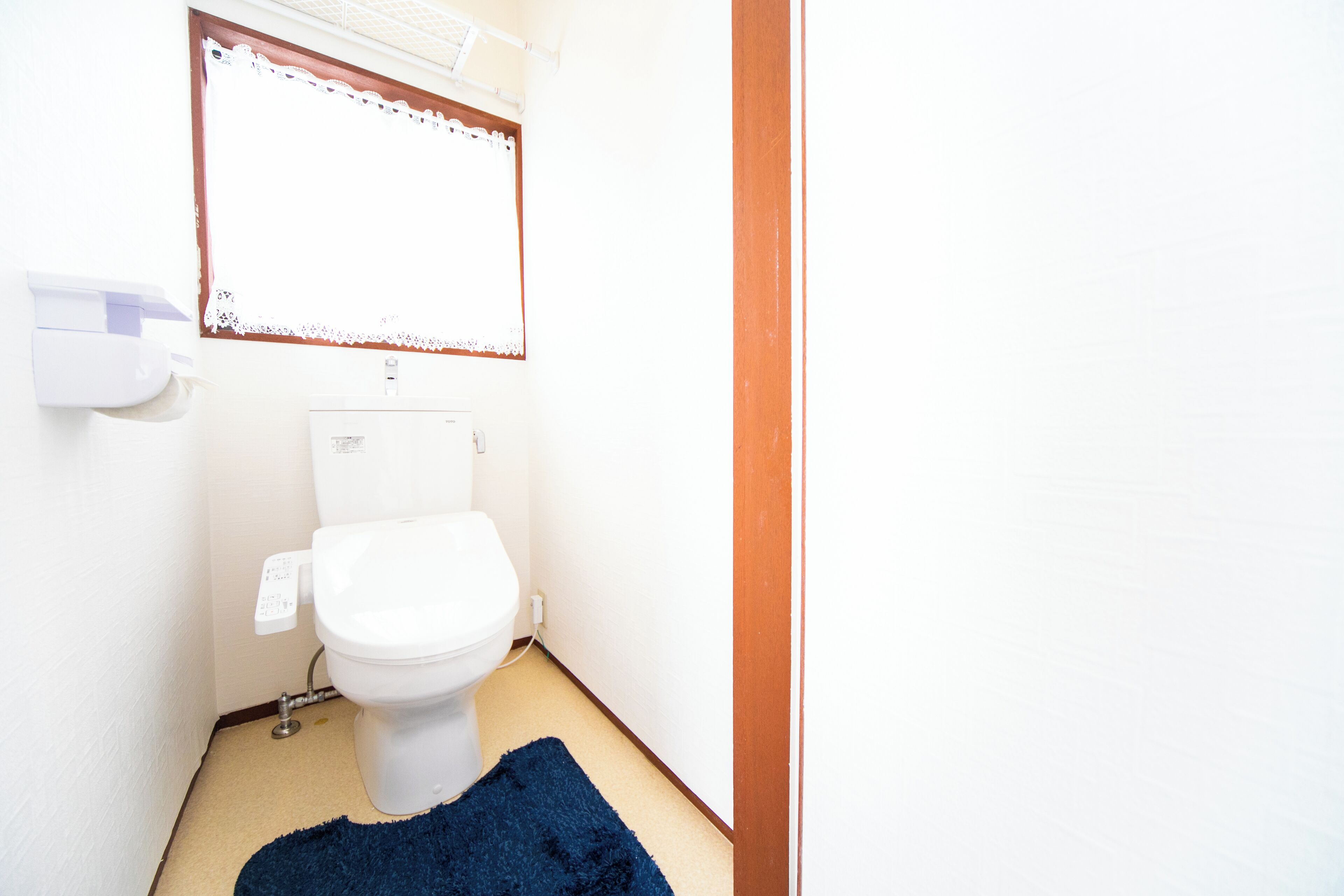 A small bathroom with a toilet, a blue rug, and a window with lace curtains.