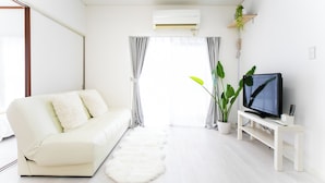 Ein modernes Wohnzimmer mit einem weißen Sofa, einem Flachbildfernseher auf einem weißen Schrank, einer großen Topfpflanze und einem flauschigen weißen Teppich.
