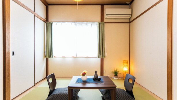 Ein traditioneller japanischer Raum mit Tatami-Böden, einem niedrigen Tisch, zwei Stühlen, einem Fenster mit grünen Vorhängen, einer Pendelleuchte und einer kleinen Topfpflanze.