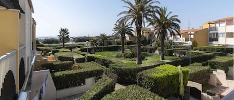 A courtyard with neatly trimmed hedges, palm trees, and a view of the sea.