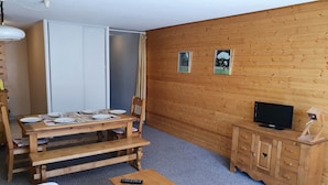 Une pièce comprenant une table à manger en bois, préparée pour quatre personnes, un téléviseur posé sur un meuble en bois, et un lit recouvert d’une couette blanche.