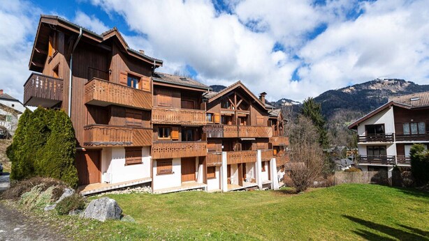 A wooden chalet with multiple balconies, a white facade, and a mountainous backdrop.