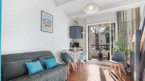 A living room with a grey sofa, a dining table with chairs, a TV, and a sliding glass door leading to an outdoor area. - Garden Retreat Near the Beach (Agde)