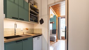 A compact kitchen with a dishwasher, a microwave, and a sink. There is a door leading to a living area with a sofa and a hanging light fixture.