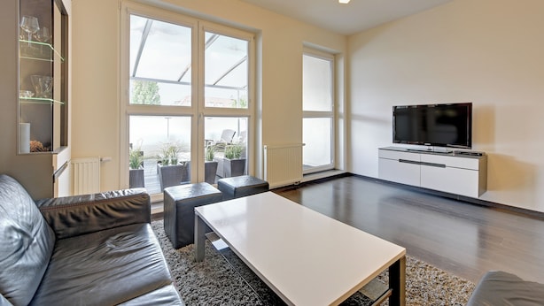 Una sala de estar moderna con un sofá, una mesa de centro y un televisor sobre un mueble bajo. Amplias ventanas ofrecen luz natural y vistas al exterior.
