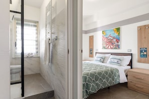 A bedroom with a bed, a wooden nightstand, a painting on the wall, and a bathroom with a shower and toilet.