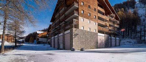 A multi-story hotel with a wooden facade and balconies, situated in a snowy landscape with trees in the background.