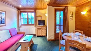 Una habitación acogedora con techo de madera, un sofá, un televisor, una mesa de comedor preparada para cuatro comensales y una ventana con cortinas.