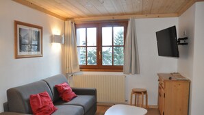 A cozy living room with a grey sofa, red cushions, a white coffee table, a wooden cabinet, and a TV mounted on the wall. - Mountain Retreat for 6 Guests (Les Deux Alpes)